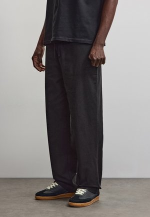 Homme debout portant un pantalon noir ample, un t-shirt noir et des baskets noires avec des lacets blancs et des semelles marron sur un sol gris.