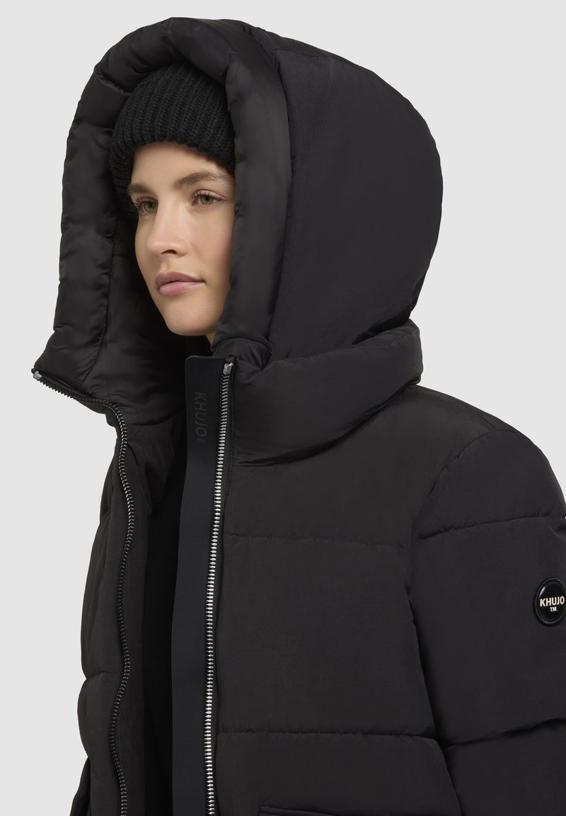 Schwarze Pufferjacke mit einer hohen, gesteppten Kapuze und einem Reißverschluss. Besitzt ein rundes Logo-Patch am linken Arm und verdeckte seitliche Taschen.