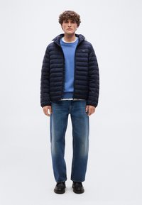 Νέος άνδρας με σγουρά μαλλιά, φορώντας μαύρο μπουφάν τύπου puffer, μπλε φούτερ, τζιν με φαρδιά πόδια και μαύρα παπούτσια, στέκεται με το πρόσωπο προς τα εμπρός.