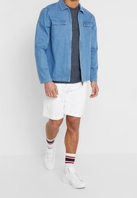 Chaqueta de mezclilla azul con dos bolsillos en el pecho, camiseta gris debajo, pantalones cortos blancos y zapatillas blancas con calcetines a rayas rojas y navy.