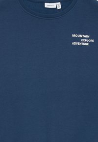 T-shirt de algodão azul-marinho com gola redonda, apresentando texto branco que diz "MOUNTAIN EXPLORE ADVENTURE" na frente. Textura suave.