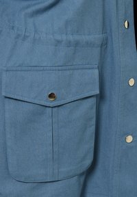 Blå tygjacka med ficka med klaff och en metalldragknapp, sydda kanter och en vertikal rad av metalldragknappar på jackans framsida.