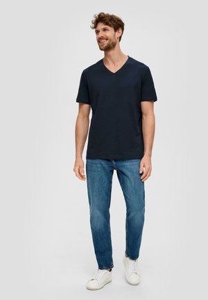T-shirt en coton bleu marine avec un col en V, associée à un jean en denim bleu clair et des baskets blanches, se tenant devant un fond neutre.