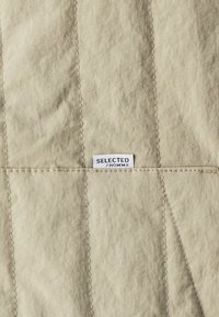 Beżowa pikowana tkanina o miękkiej fakturze z ukośnym przeszyciem. Przymocowana jest biała metka z napisem "SELECTED HOMME".
