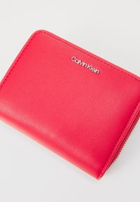 Röd konstläderplånbok med en slät yta, prydd med en silverfärgad Calvin Klein-logotyp, dragkedjestängning och rundade hörn.