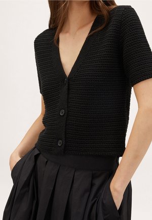 Donna che indossa un cardigan nero a maniche corte con bottoni e una gonna nera a pieghe, con le mani nelle tasche della gonna, in piedi contro uno sfondo bianco.