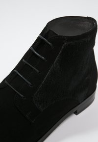 Bottine en daim noir avec une finition lisse, une texture subtile, cinq œillets avec des lacets noirs, et une semelle noire plate.