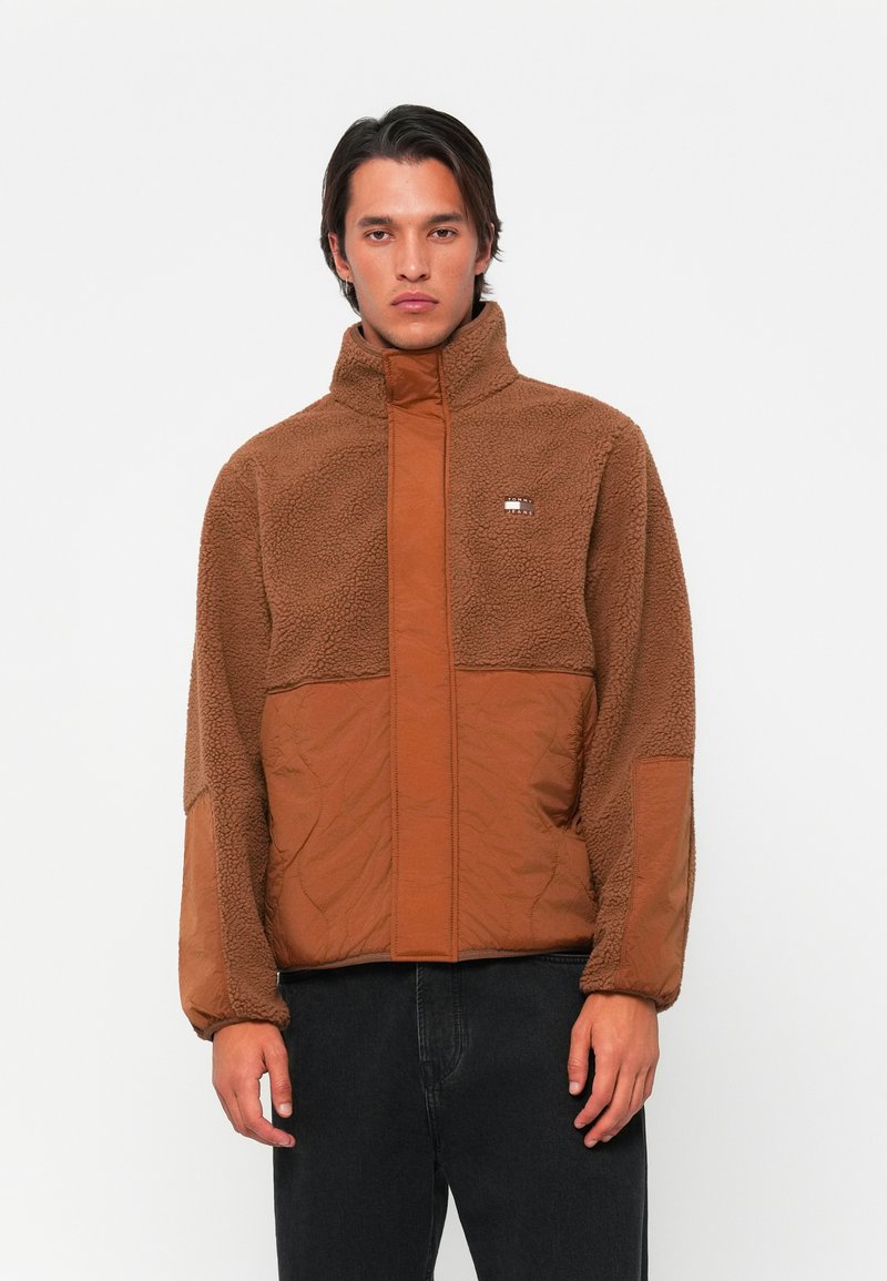 Brown fleece jacket with a contrasting brown panel, high collar, zip front, and textured sleeve accents. Features logo on chest area.