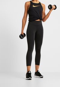 Top sportivo nero con logo Nike dorato, abbinato a leggings neri e scarpe da ginnastica nere, mentre tiene i manubri in entrambe le mani.