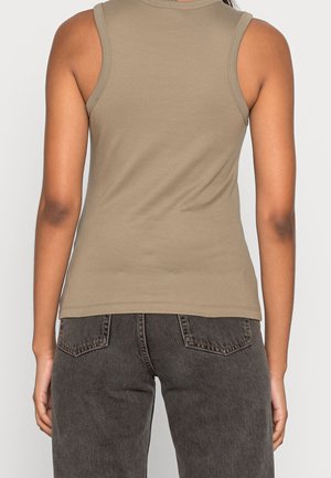 Person, die ein beiges ärmelloses Oberteil und dunkelgraue Jeans trägt, von hinten vor einem einfarbigen hellen Hintergrund gezeigt.