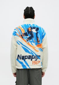 Fliiskangast jakk, millel on kreemjas alus koos sinise, oranži ja tumesinise abstraktsete mustritega; seljal suur "Napapijri 1987" logo.