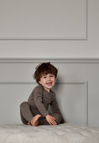 Body marrone a maniche lunghe indossato da un bambino con capelli scompigliati, seduto su una superficie testurizzata di colore chiaro contro una parete dai toni smorzati.