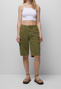 Shorts cargo vert olive avec six poches, ourlet retroussé et tissu en coton léger extensible. Associés à un haut court blanc et des sandales beiges.