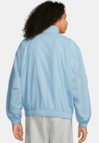 Giacca oversized blu chiaro con colletto alto, polsini e vita elasticizzati. Presenta una superficie liscia e elementi di design minimi.