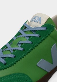 Baskets vertes avec une tige synthétique lisse, des lacets bleu clair, une languette blanche contrastante et une semelle en caoutchouc marron. Présentent un logo bien visible.