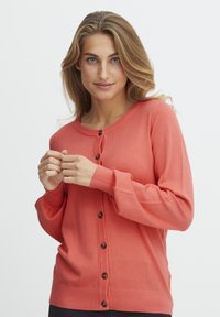Cardigan en tricot corail avec un col rond, des manches longues, des poignets côtelés et cinq boutons marron sur le devant. Texture douce.