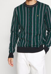 Suéter verde con estampado, de cuello redondo, con diseños geométricos blancos y una banda oscura en el dobladillo y los puños. Detalle de logo bordado visible.