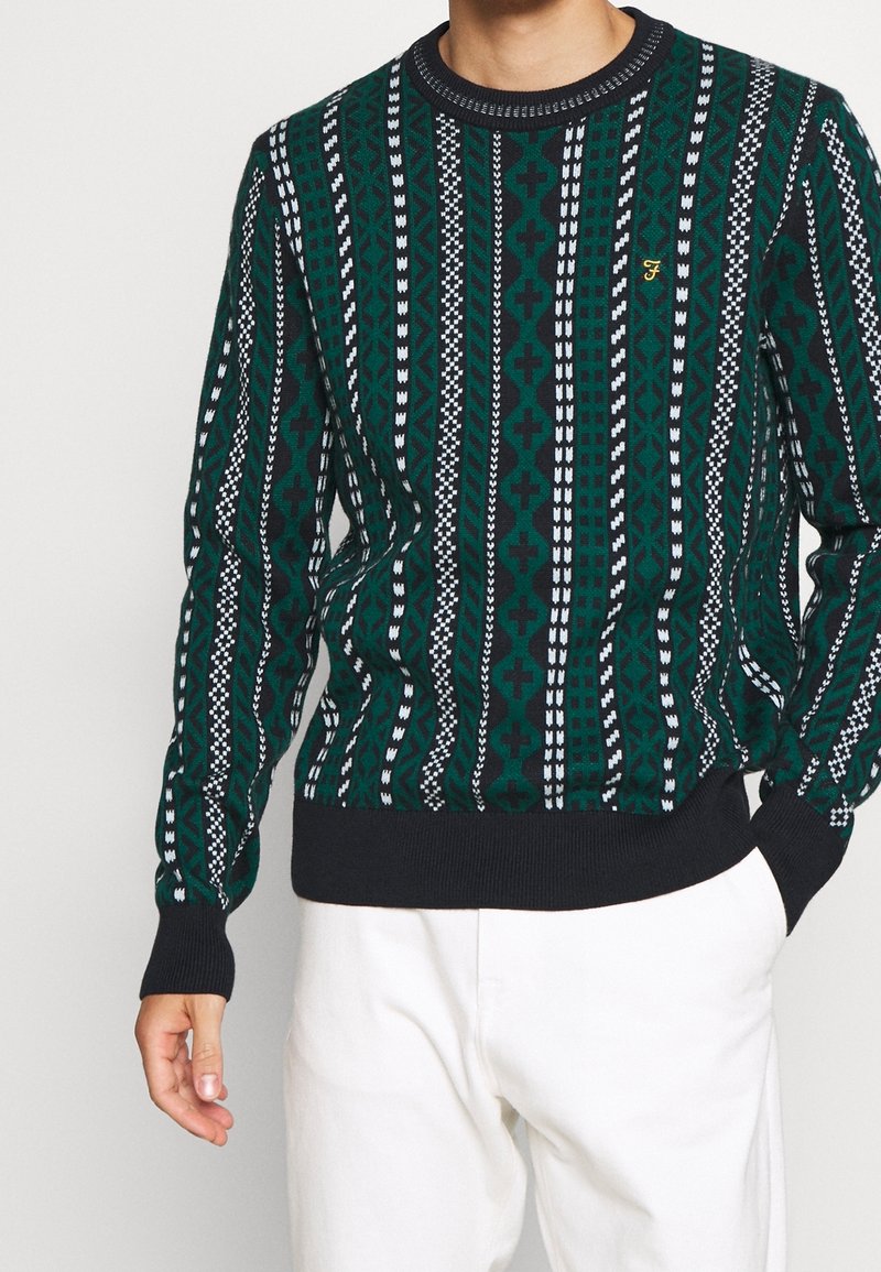 Suéter verde con estampado, de cuello redondo, con diseños geométricos blancos y una banda oscura en el dobladillo y los puños. Detalle de logo bordado visible.