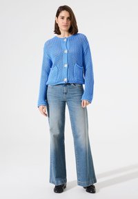 Blauer gestrickter Cardigan mit einem strukturierten Muster, ausgestattet mit zwei vorderen Taschen und drei Knöpfen. Tragen Sie ihn zu hellblauen Schlagjeans.