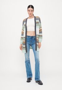 Πλεκτό cardigan με πολύχρωμες ρίγες σε πράσινο, καφέ και λευκό. Διαθέτει πλατύ ανοιχτό μπροστά, ριμπ μανσέτες και χαλαρή γραμμή. Συνδυάζεται με μπλε τζιν.