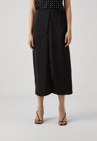 Jupe midi en satin noir avec une texture lisse, présentant une couture frontale et une silhouette légèrement évasée, assortie de talons sandales noirs minimalistes.