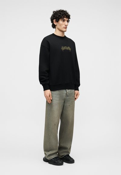 Άνδρας που στέκεται φορώντας μαύρο φούτερ με μικρό κίτρινο γραφικό, φαρδιά ξεθωριασμένα γκρι τζιν και μαύρα αθλητικά παπούτσια, μπροστά σε απλό φόντο.