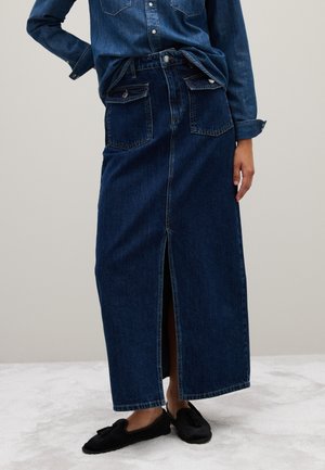 Denim maxi skirt in dark blue with a front slit. Features two front pockets and a waistband with belt loops. Paired with black slippers.