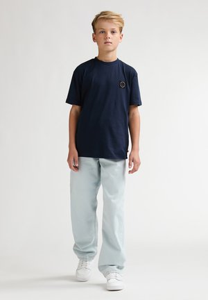 Blond jongetje staand, draagt een marineblauw T-shirt, lichtgrijze wijde broek en witte sneakers tegen een effen lichte achtergrond.