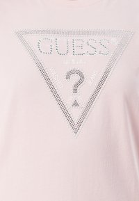 Camiseta de manga corta rosa claro con un diseño de strass triangular que presenta el texto "GUESS U.S.A. WASHED JEANS" en plata.