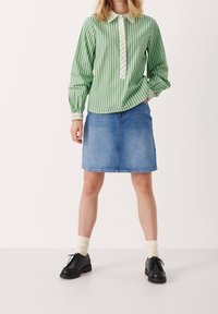 Zeleno-bíle pruhovaná košile s dlouhým rukávem a klasickým límcem, doplněná modrou džínovou sukní. Černé boty a světlé ponožky dotvářejí outfit.