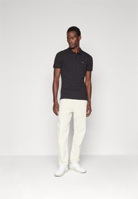 Camisa polo negra de manga corta, corte ajustado, con logo bordado en el pecho, combinada con pantalones chinos beige claro y zapatillas blancas.