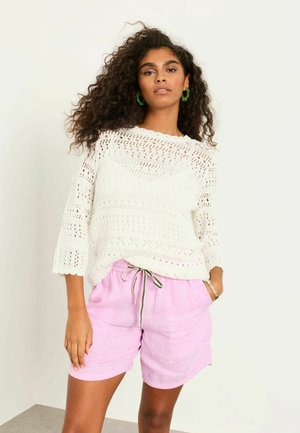 Frau mit lockigem Haar, die ein weißes Häkeloberteil, hellrosa Shorts mit Kordelzug, grüne Creolen und ein Armband trägt, steht mit einer Hand in der Tasche.