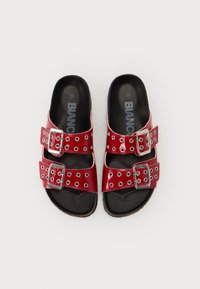 Sandales en faux cuir rouge avec deux brides ajustables ornées de boucles en argent et d'œillets décoratifs, posées sur une semelle noire rembourrée.