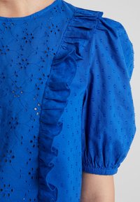 Niebieska bluzka z haftowanymi kwiatowymi wzorami, falbaną z przodu oraz krótkimi bufkami z elastycznymi mankietami.