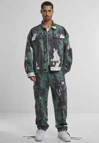 Chaqueta de mezclilla con estampado y pantalones cargo en verde, púrpura y negro, que presentan formas geométricas, múltiples bolsillos y acentos en blanco contrastantes.