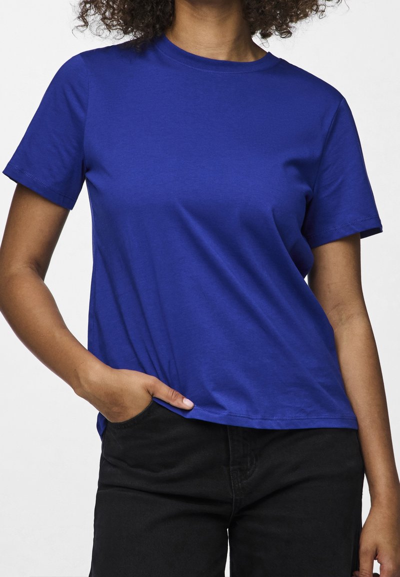 Persona che indossa una maglietta semplice di colore blu royal a maniche corte e pantaloni neri, con una mano appoggiata in modo casuale in tasca.