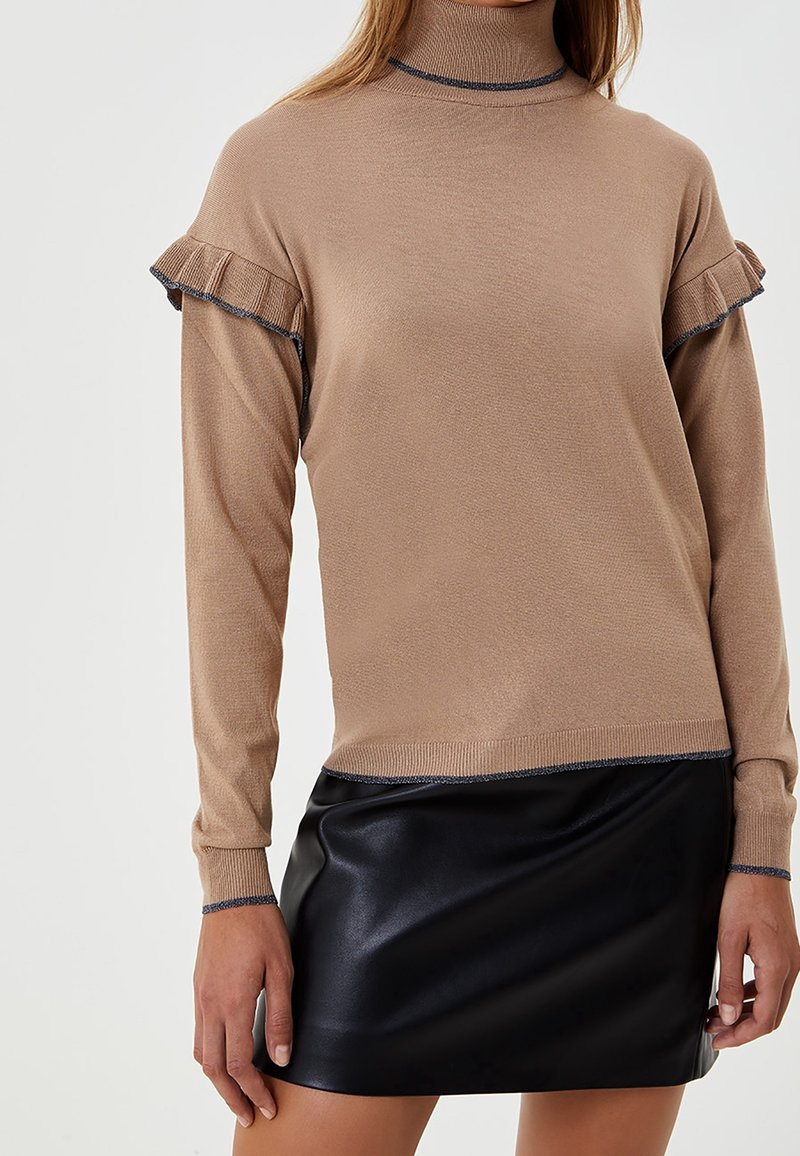 Suéter de cuello alto beige con acentos de volantes y ribete oscuro en las mangas, combinado con una falda mini de cuero sintético negra. Tejido suave y texturizado.