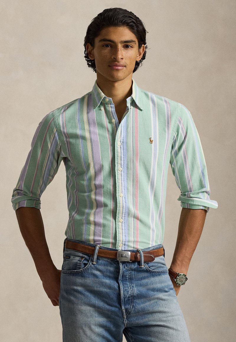 Mladi muškarac nosi košulju na pruge u pastelnoplavoj, lavandinoj i bijeloj boji s podvrnutim rukavima, plave traperice, smeđi remen i ručni sat.