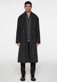 Cappotto lungo doppiopetto in misto lana grigio scuro, con grandi rever, bottoni neri e tasche laterali. Indossato sopra un maglione in maglia grigio.