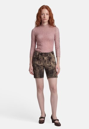 Roodharige vrouw die een roze kanten top met lange mouwen, donkerbruine leren shorts met borduursel en Mary Jane schoenen met hakken draagt, rechtop staand.