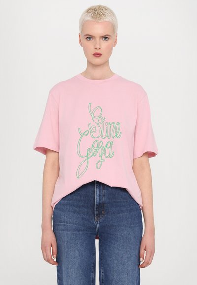 T-shirt de algodão rosa com mangas curtas e um corte largo, apresentando o texto em verde em letra cursiva "Sine Yoga" ao centro. Usado com jeans de ganga escuros.