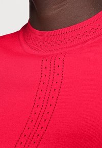 Pull tricot rouge avec un col montant, un détail perforé complexe sur l'épaule et une texture lisse et élégante.