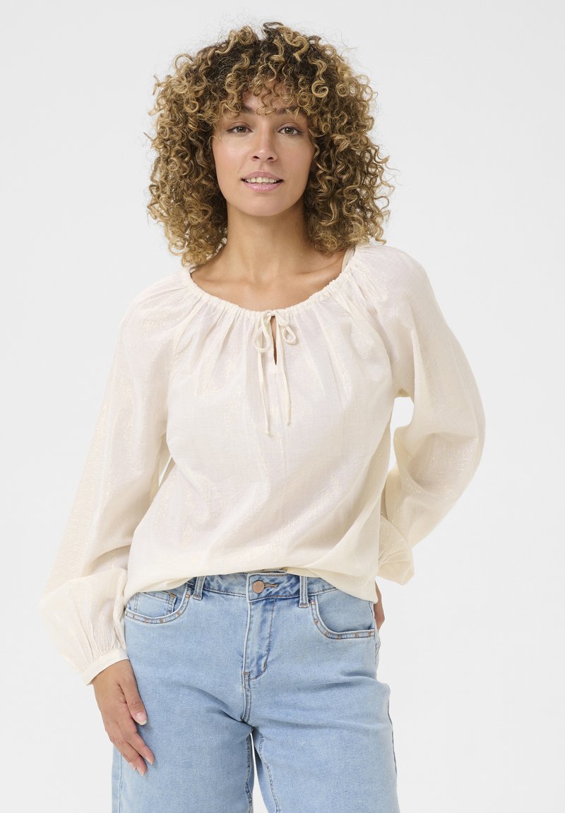 Light-colored, textured blouse with a rounded neckline, drawstring detail, and billowing sleeves, paired with light blue jeans.