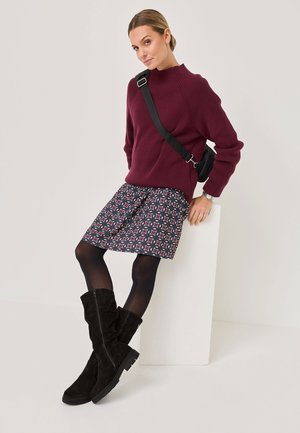 Bordeauxfarbener gerippter Pullover, gemusterter marineblauer Rock mit roten und weißen Akzenten, schwarze kniehohe Wildlederstiefel und eine schwarze Umhängetasche.