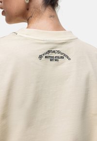 Crème-kleurige sweatshirt met een relaxte pasvorm, voorzien van een zwart geborduurd merklogo op de achterkant en een gladde, zachte textuur.
