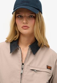 Helle beigefarbene Jacke mit einem dunklen Cordkragen, Reißverschluss und einer Fronttasche. Dunkelblaue Kappe mit dezentem Logo. Glatte Stofftextur.