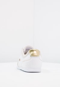 Bílá teniska s texturovaným svrchním materiálem, zlatým akcentem na patě a minimalistickým designem. Hladká podrážka s kulatým tvarem.