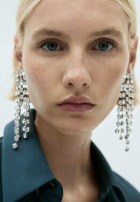 Sparkling silver statement earrings with cascading geometric shapes, hanging from the model's ears, set against a neutral background.