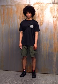 Maglietta nera con logo, pantaloni cargo verdi e stivali neri. Il modello è in piedi su una superficie grigia testurizzata contro uno sfondo color ruggine.