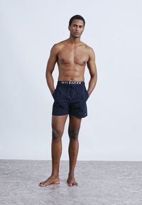 Μαύρο μαγιό σορτς με κορδόνι στη μέση και λευκή λωρίδα με λογότυπο, κατασκευασμένο από ελαφρύ ύφασμα· το μοντέλο στέκεται ξυπόλυτο μπροστά σε ουδέτερο φόντο.